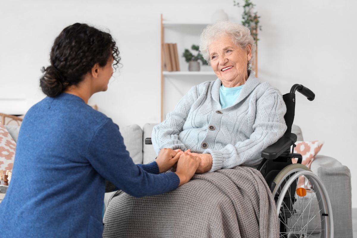 A carer holds an elderly woman's hands as she sits in a wheelchair.