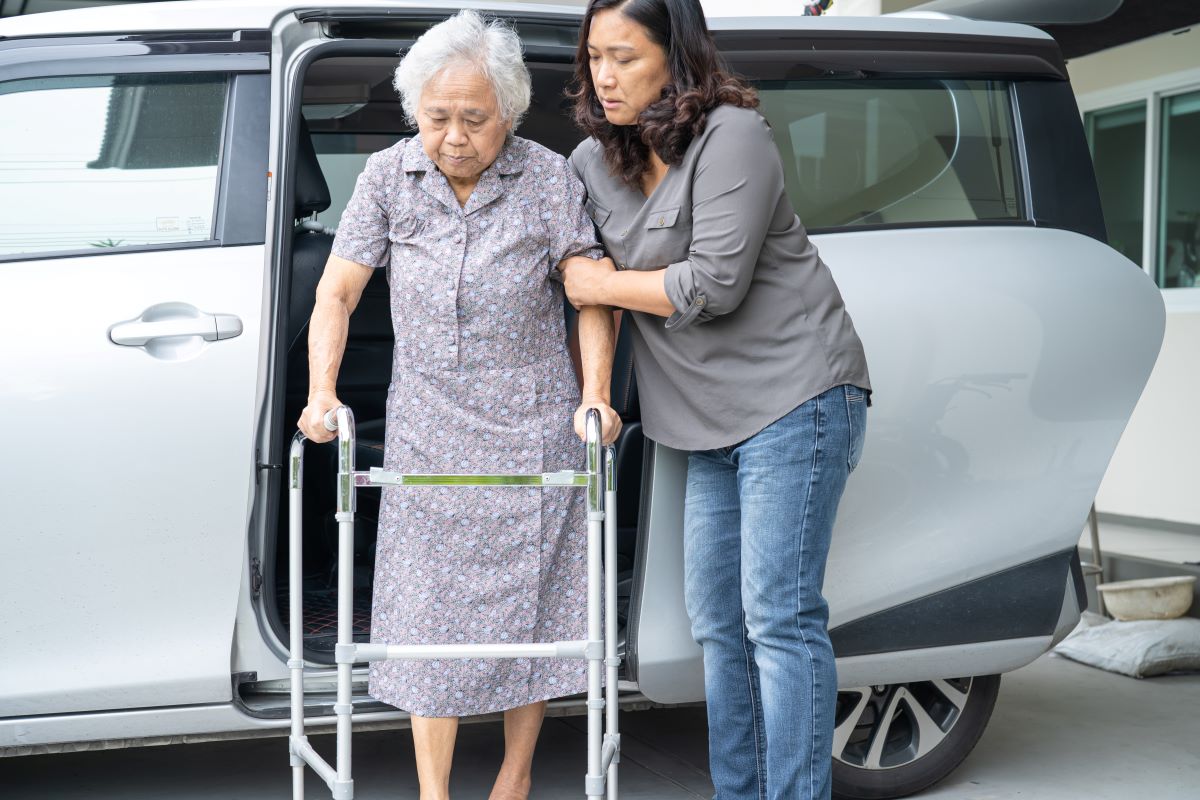 A support worker helps an elderly woman out of a vehicle.