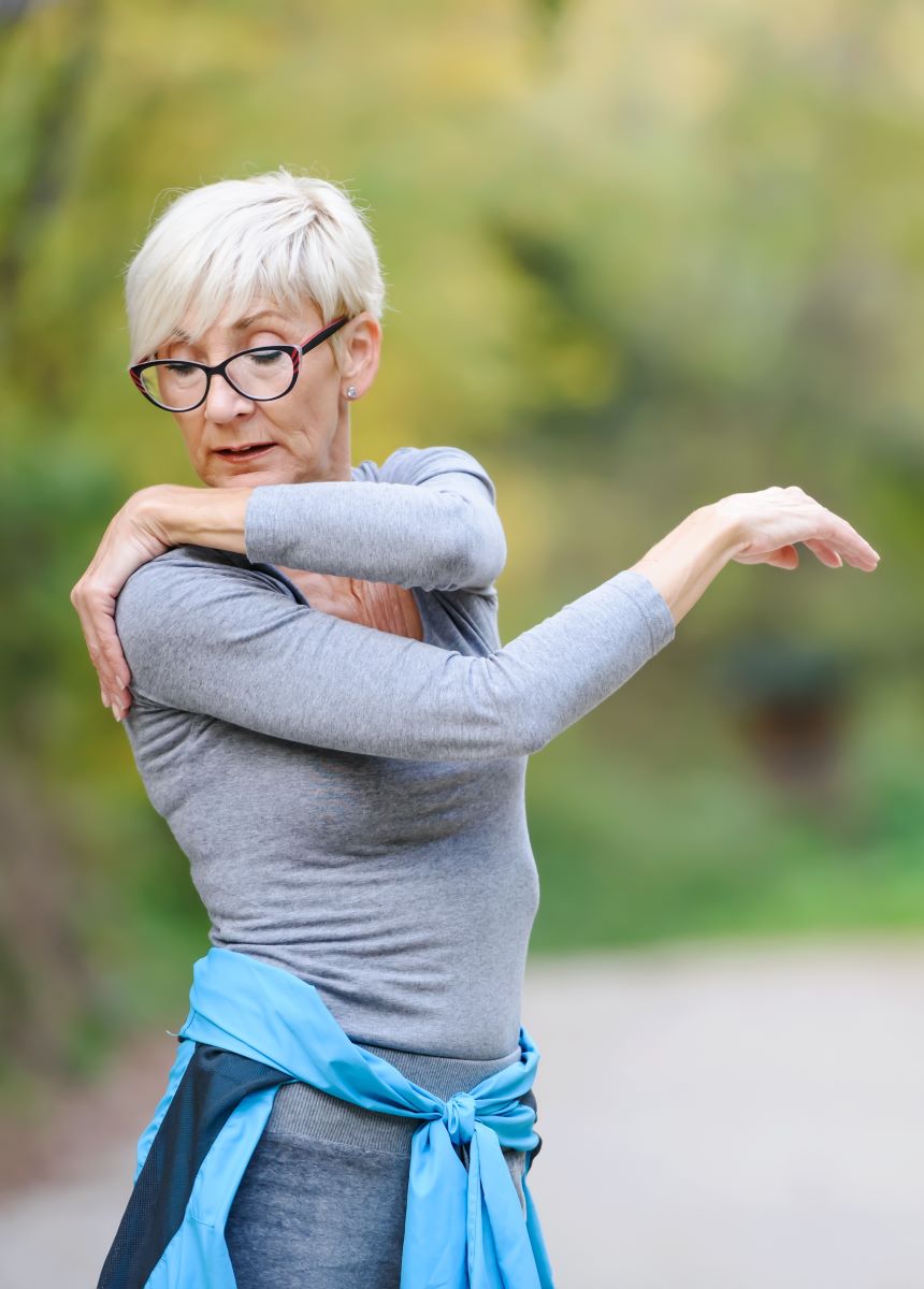 An elderly woman holds her sore shoulder while exercising outdoors.