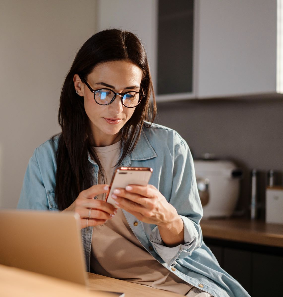 A woman sits at her home workstation and uses her phone.