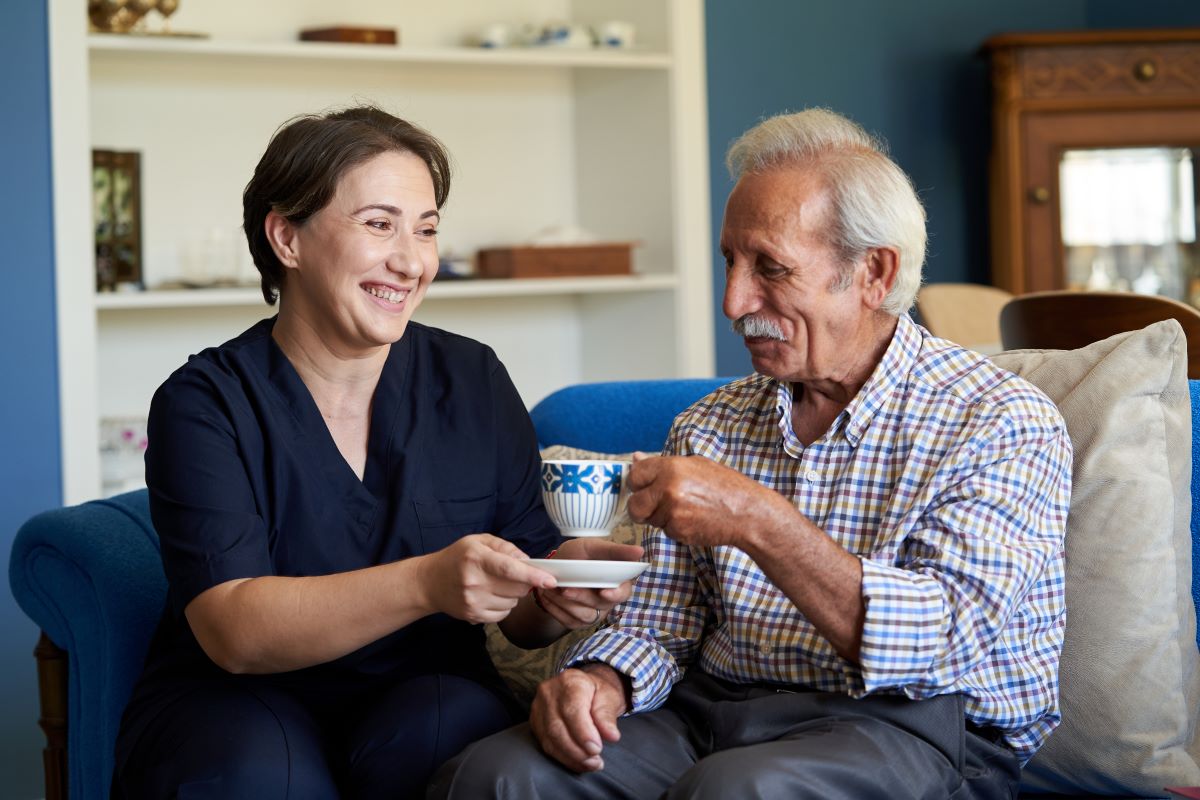 A carer hands an elderly man a cup of tea.